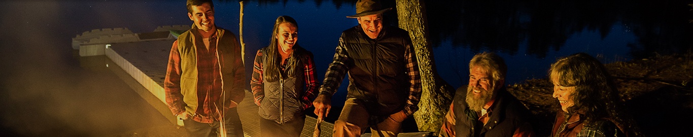 A family gathers around a campfire with a dock in the background