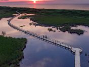 Wide shot of a long floating bridge going through calm wetlands, backdropped by a coastal sunset