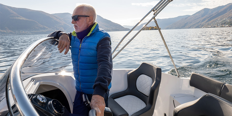 Older man in a blue jacket driving a boat over a calm lake during winter